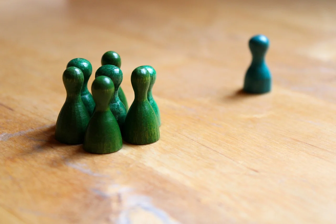 Seven green board game figures in a close circle on a wooden table and one blue board game figure out of focus, at some distance from the green group.
